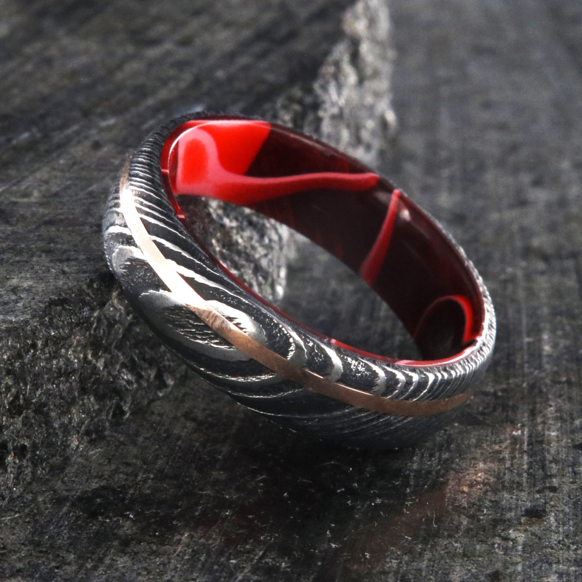 6mm wide black Damascus steel ring with a thin off-centered rose gold inlay and dark and bright red swirled acrylic sleeve