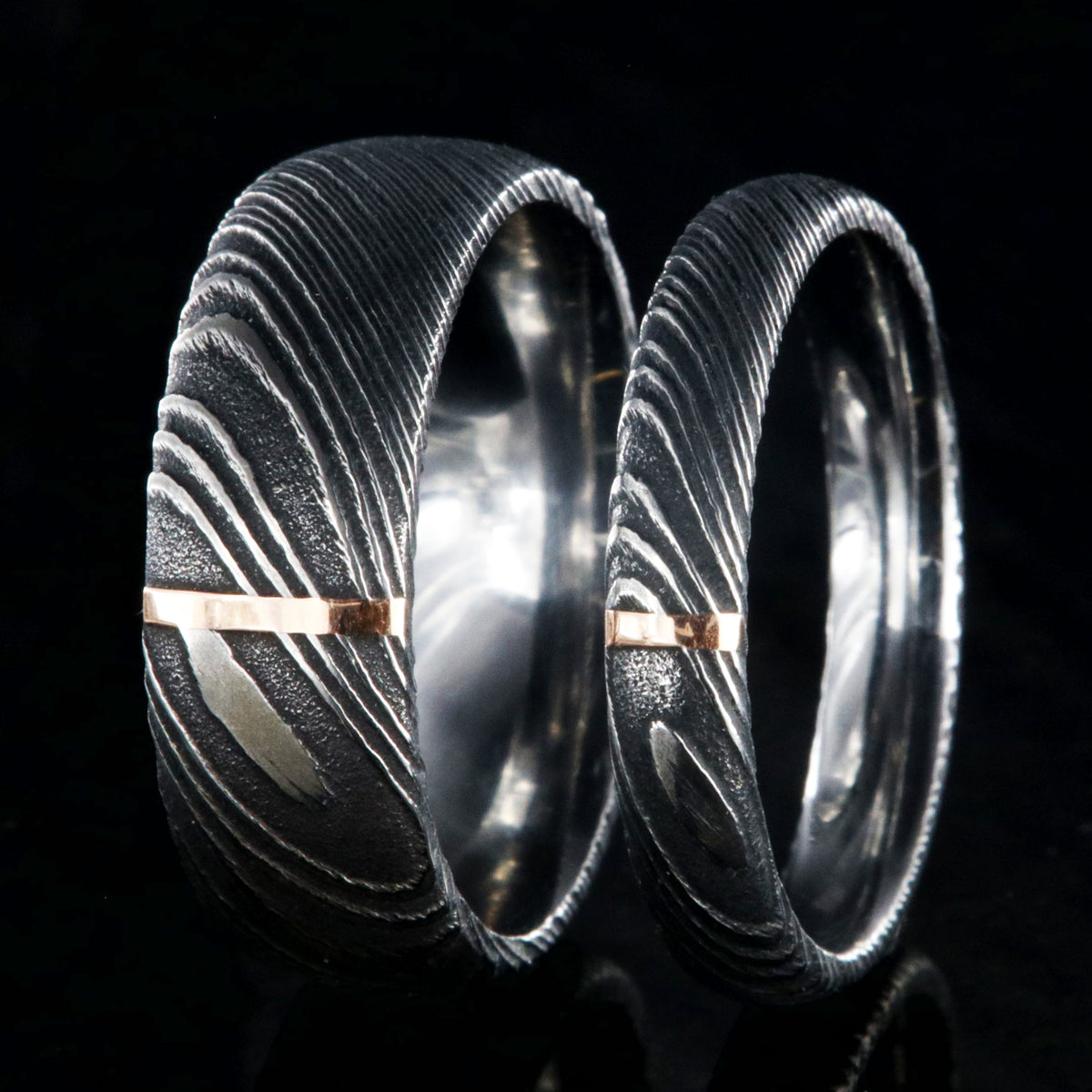 A matching black Damascus steel wedding band set with 8mm and 4mm wide rings and a single vertical rose gold inlay