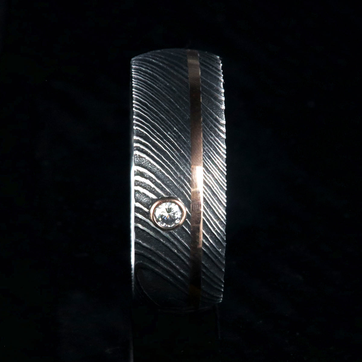 8mm wide black Damascus steel ring with rounded profile, polished inside, an off-center rose gold inlay and 3mm round bezel set diamond