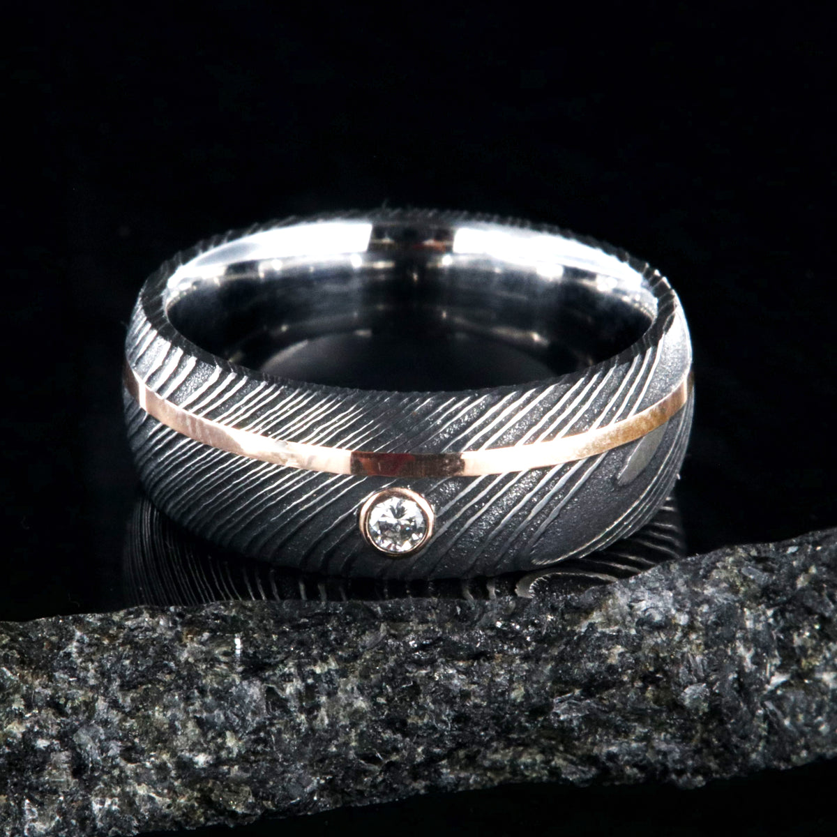 8mm wide black Damascus steel ring with rounded profile, polished inside, an off-center rose gold inlay and 3mm round bezel set diamond