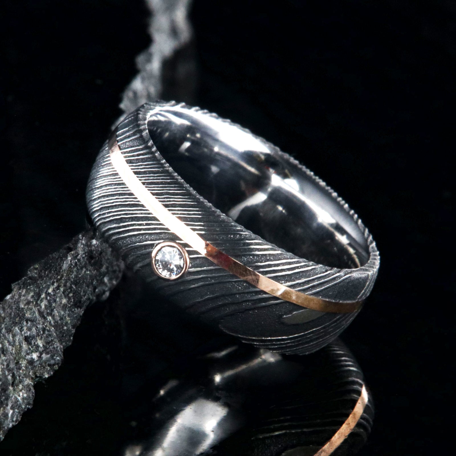 8mm wide black Damascus steel ring with rounded profile, polished inside, an off-center rose gold inlay and 3mm round bezel set diamond