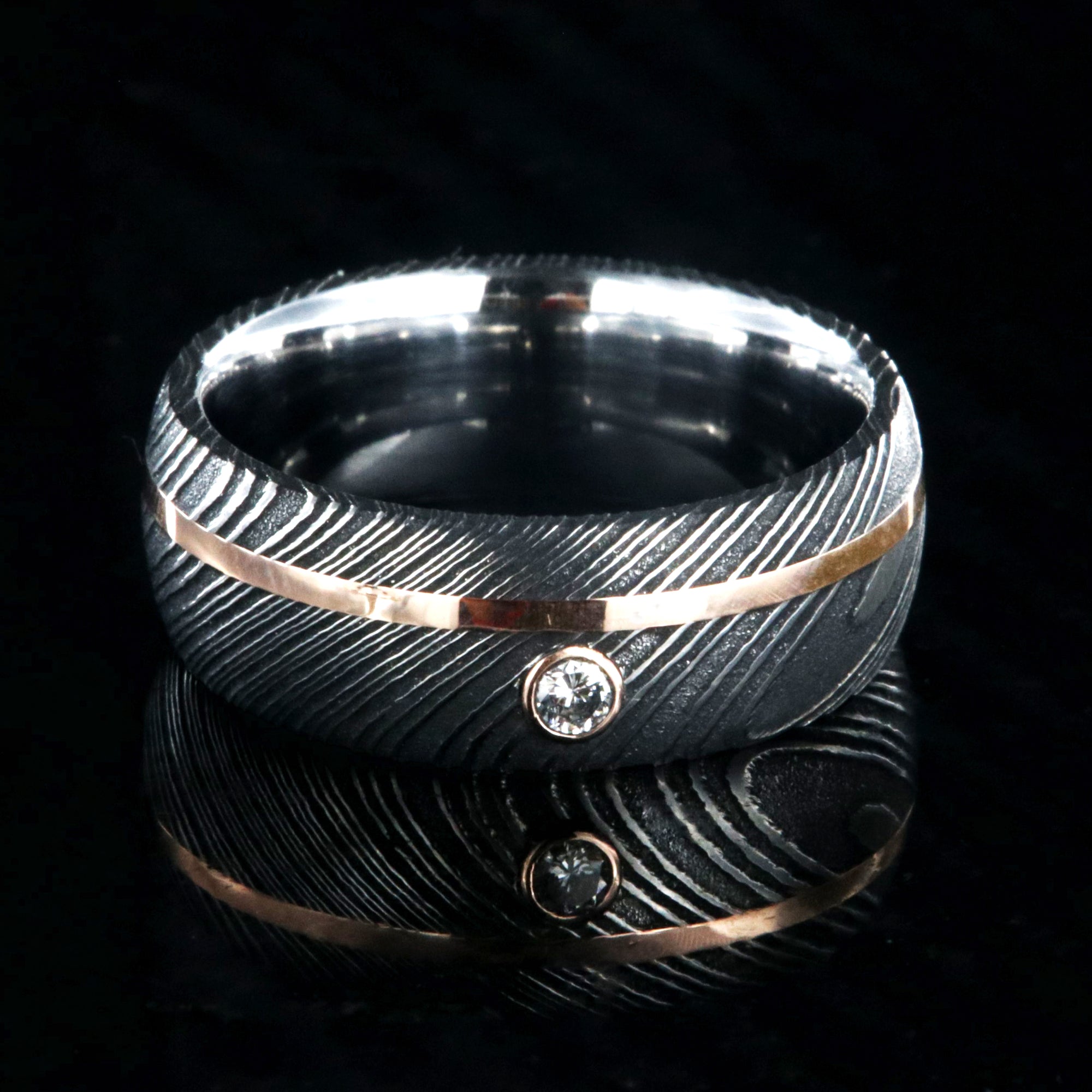 8mm wide black Damascus steel ring with rounded profile, polished inside, an off-center rose gold inlay and 3mm round bezel set diamond