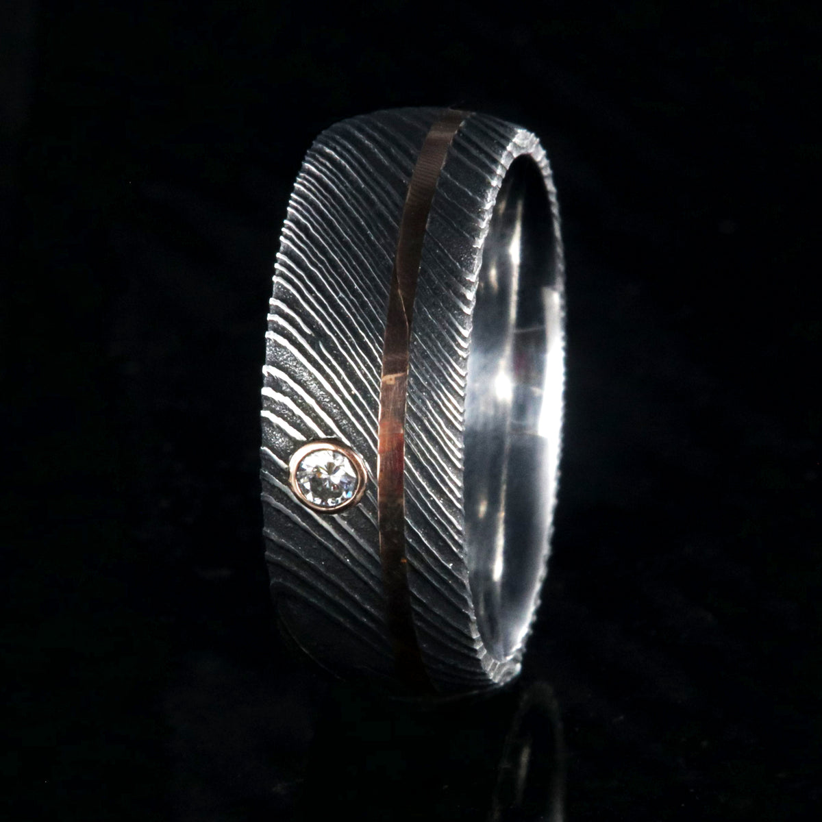8mm wide black Damascus steel ring with rounded profile, polished inside, an off-center rose gold inlay and 3mm round bezel set diamond