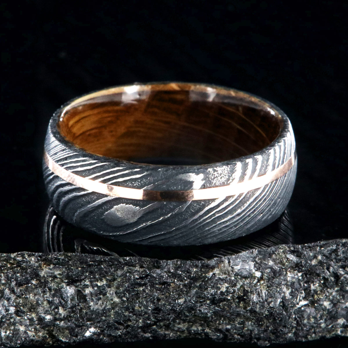 8mm wide black Damascus steel wedding ring with an off-centered rose gold inlay and a Jack Daniel&#39;s whiskey barrel sleeve