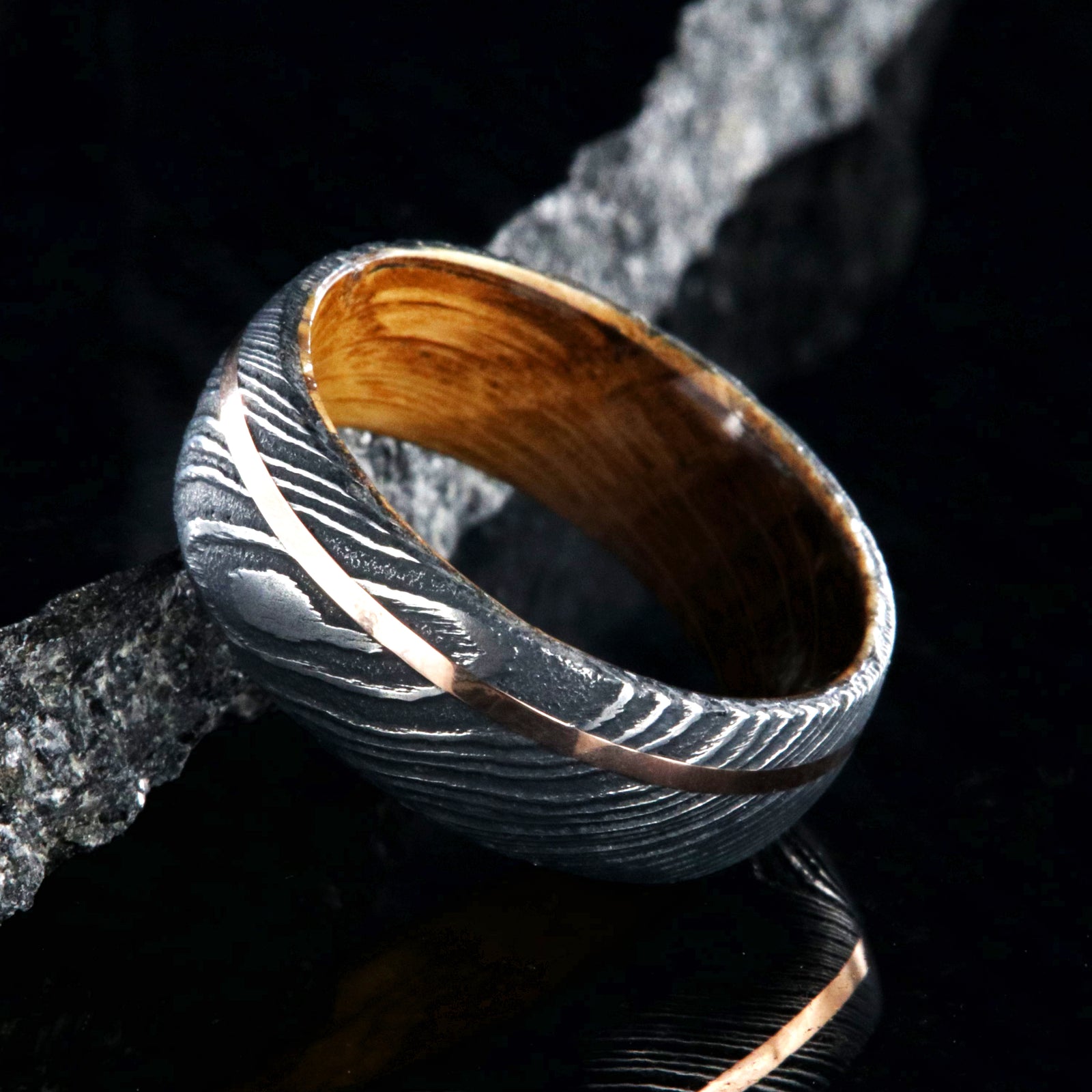 8mm wide black Damascus steel wedding ring with an off-centered rose gold inlay and a Jack Daniel's whiskey barrel sleeve