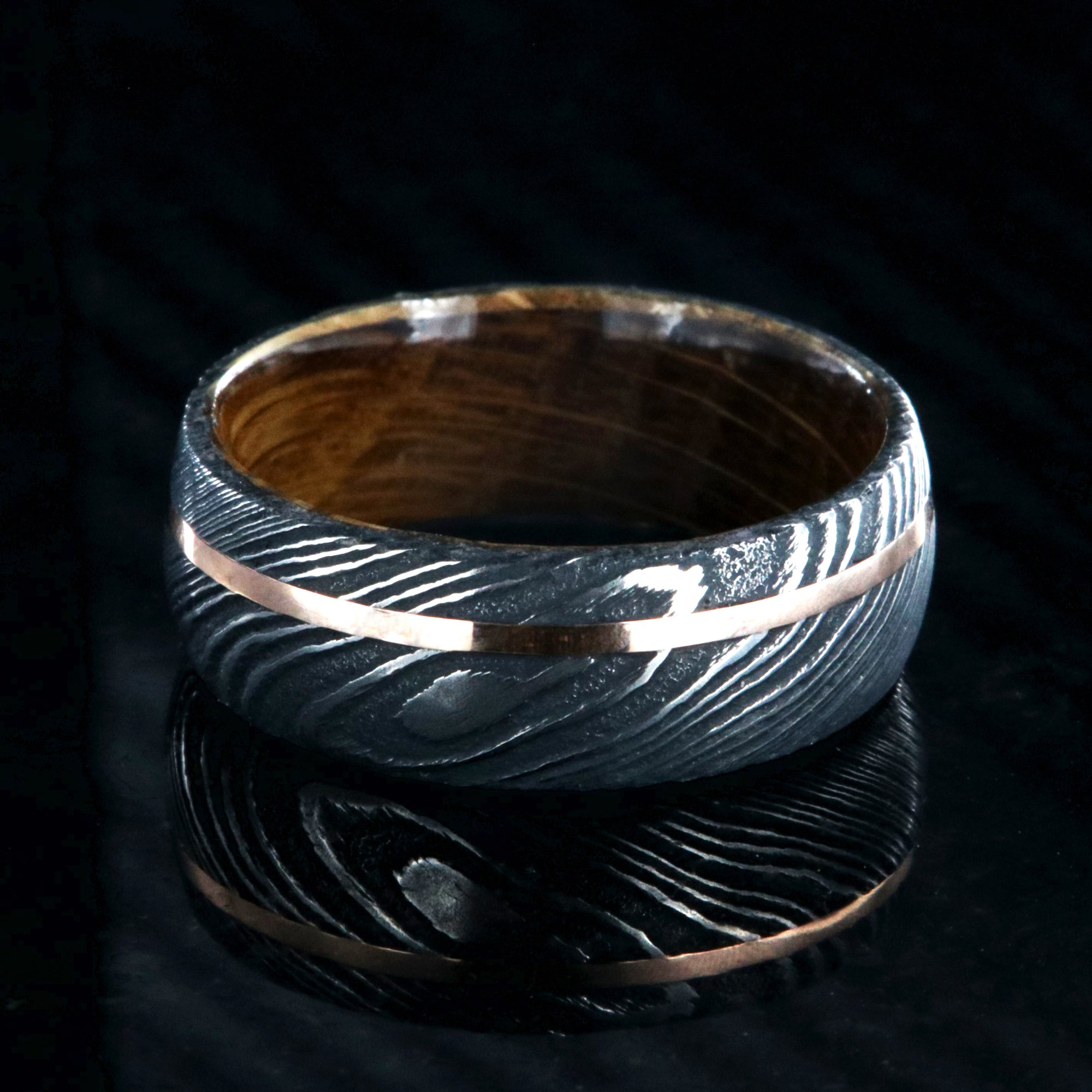 8mm wide black Damascus steel wedding ring with an off-centered rose gold inlay and a Jack Daniel's whiskey barrel sleeve