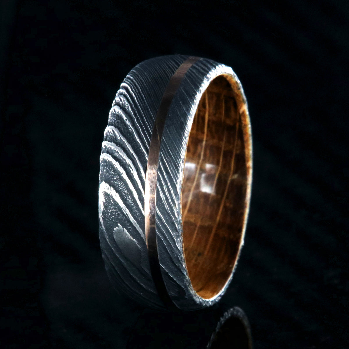 8mm wide black Damascus steel wedding ring with an off-centered rose gold inlay and a Jack Daniel&#39;s whiskey barrel sleeve