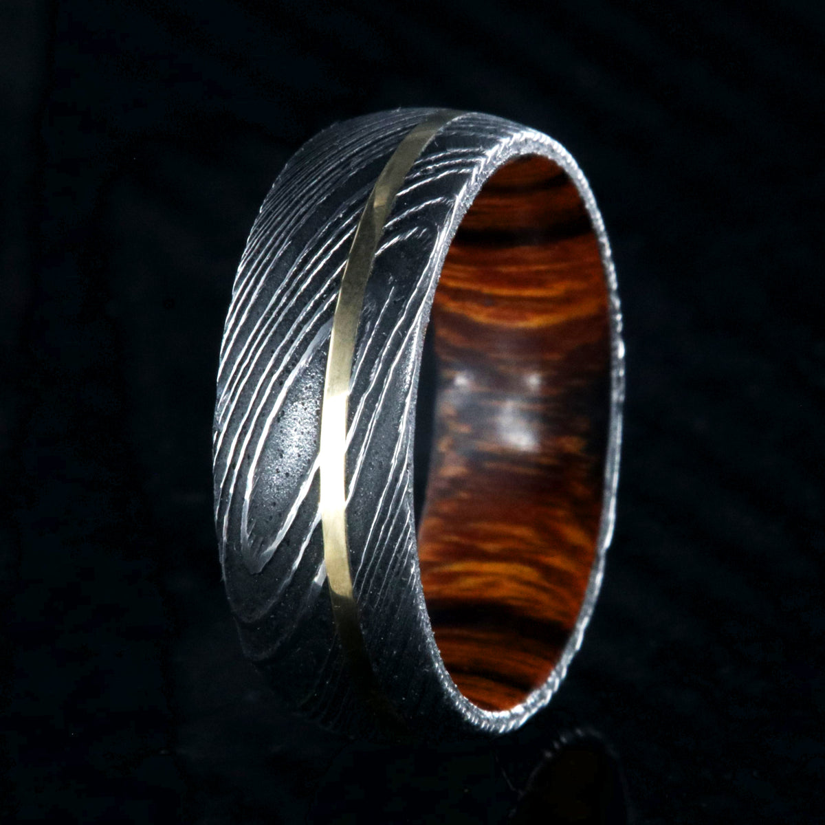 8mm wide black Damascus steel wedding band with rounded profile, an off-center yellow gold inlay, and Arizona ironwood sleeve