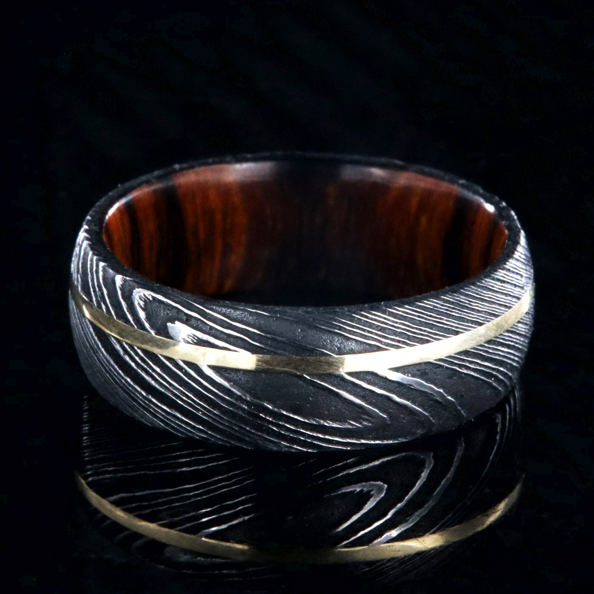 8mm wide black Damascus steel wedding band with rounded profile, an off-center yellow gold inlay, and Arizona ironwood sleeve