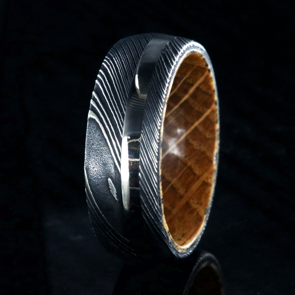8mm wide mens ring with black Damascus steel and a wide gold off-center inlay and a Jack Daniels whiskey barrel sleeve