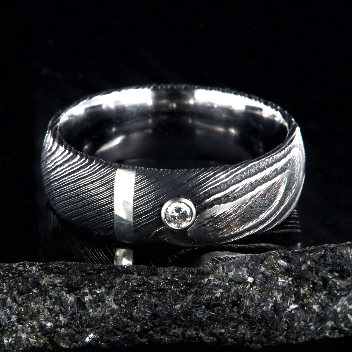 8mm wide black Damascus steel ring with polished inside, rounded profile, a vertical sterling silver inlay, and bezel set diamond