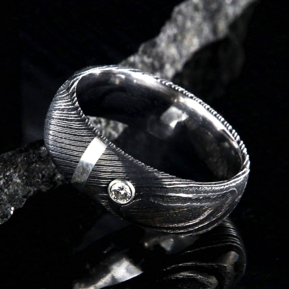 8mm wide black Damascus steel ring with polished inside, rounded profile, a vertical sterling silver inlay, and bezel set diamond