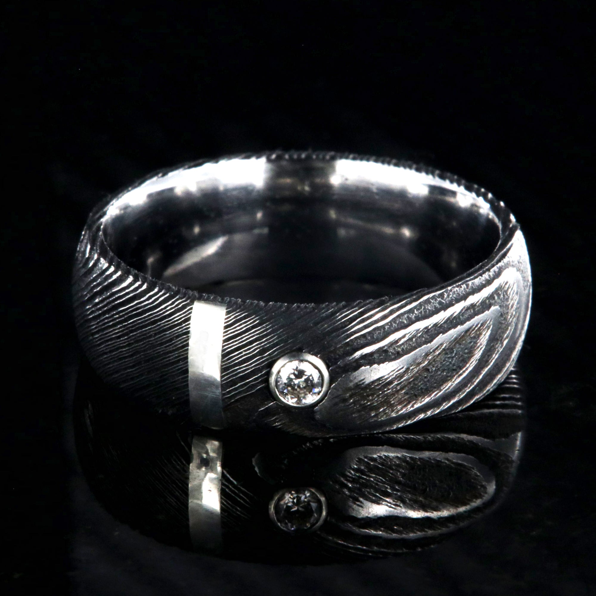 8mm wide black Damascus steel ring with polished inside, rounded profile, a vertical sterling silver inlay, and bezel set diamond