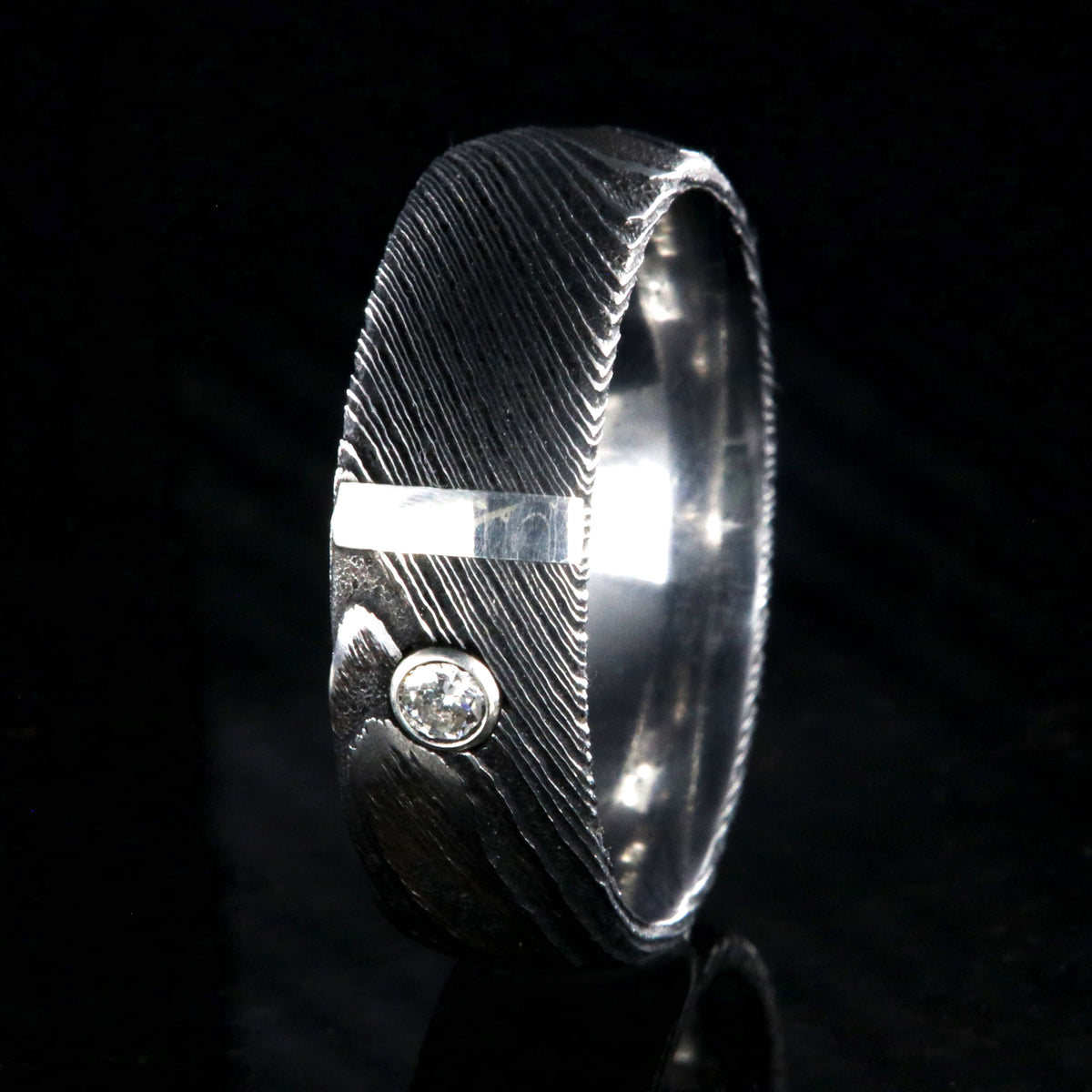 8mm wide black Damascus steel ring with polished inside, rounded profile, a vertical sterling silver inlay, and bezel set diamond