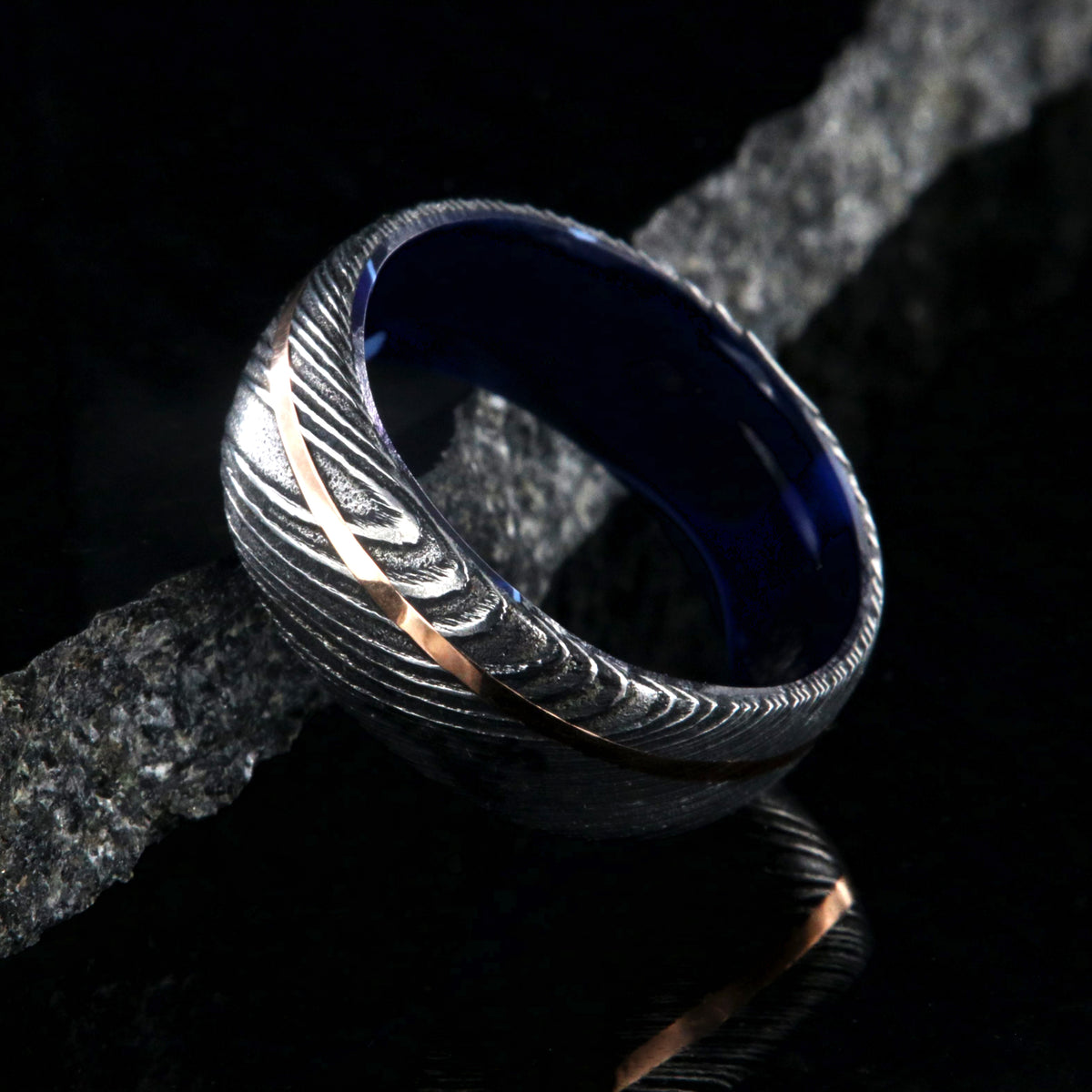 8mm wide black Damascus steel ring with an off-center rose gold inlay and dark blue acrylic sleeve