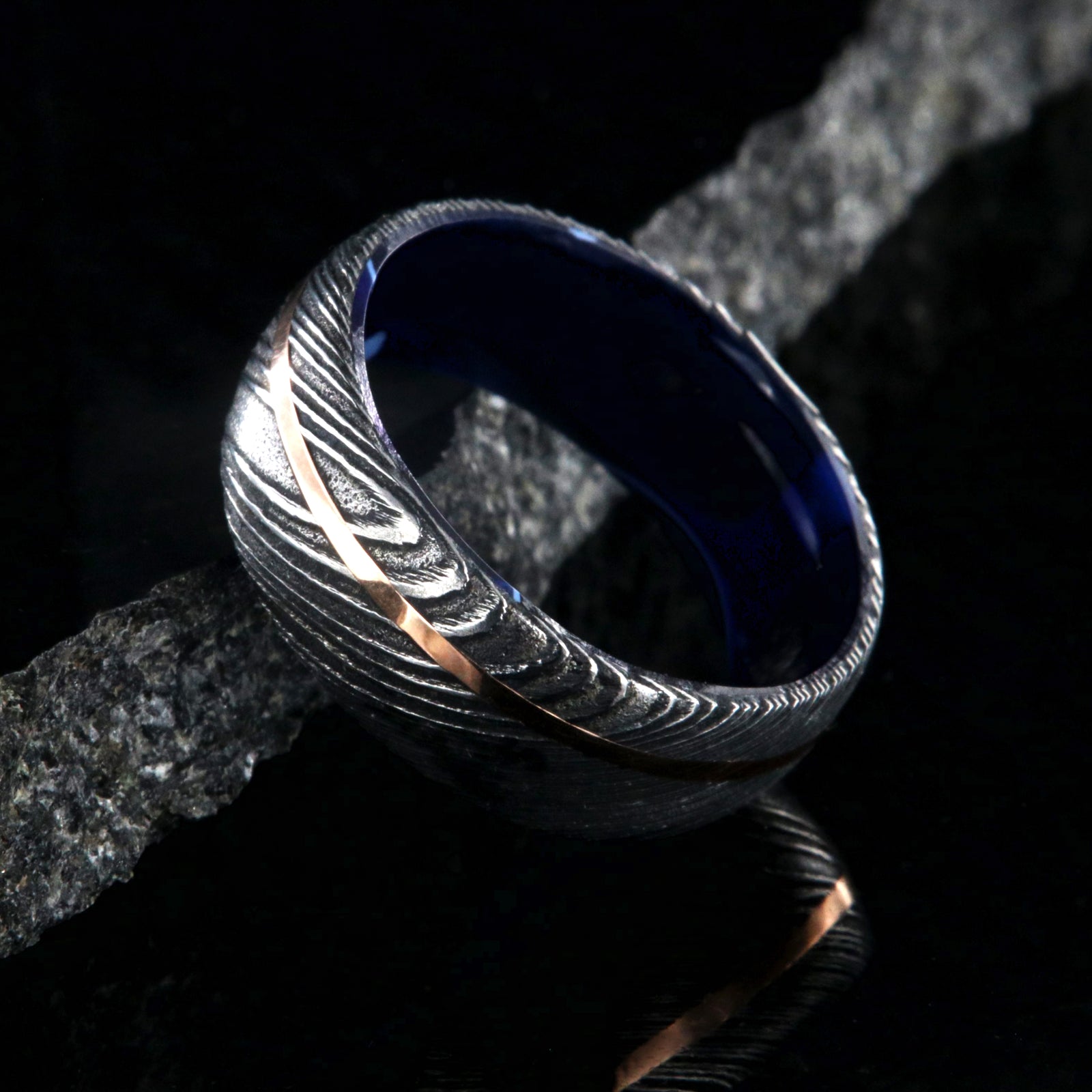 8mm wide black Damascus steel ring with an off-center rose gold inlay and dark blue acrylic sleeve