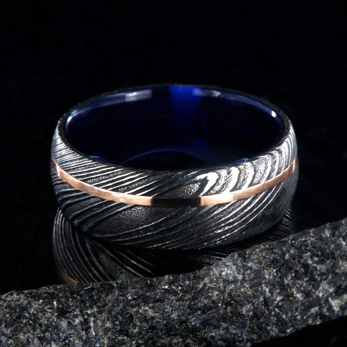 8mm wide black Damascus steel ring with an off-center rose gold inlay and dark blue acrylic sleeve