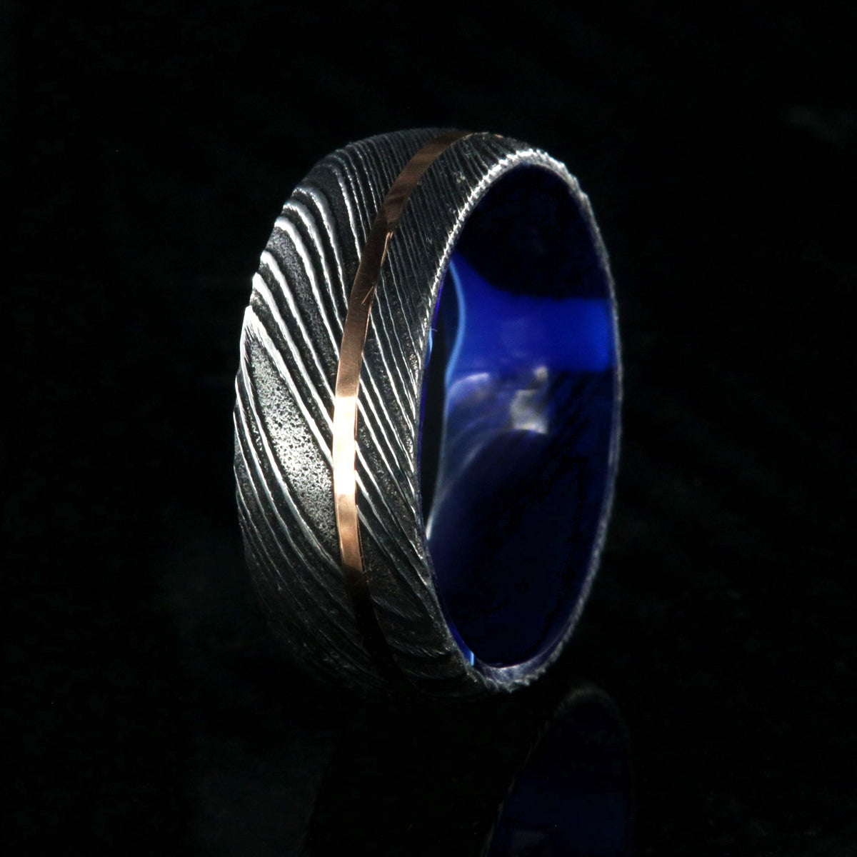 8mm wide black Damascus steel ring with an off-center rose gold inlay and dark blue acrylic sleeve
