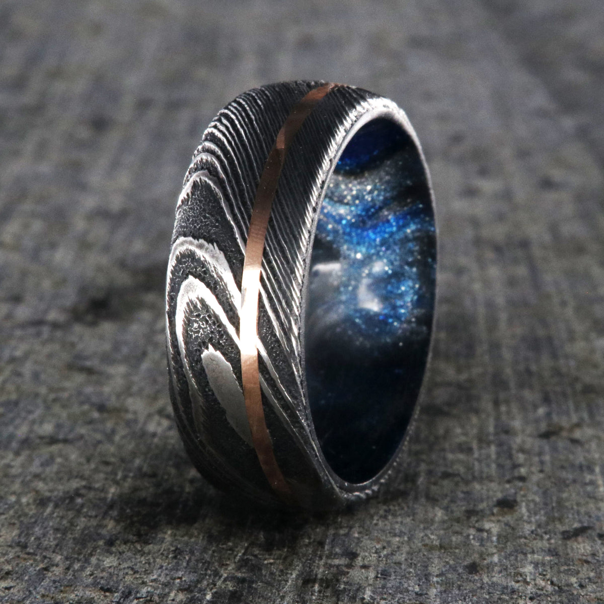 8mm wide Damascus steel wedding band with a thin rose gold off-center inlay and glittering blue and silver sleeve