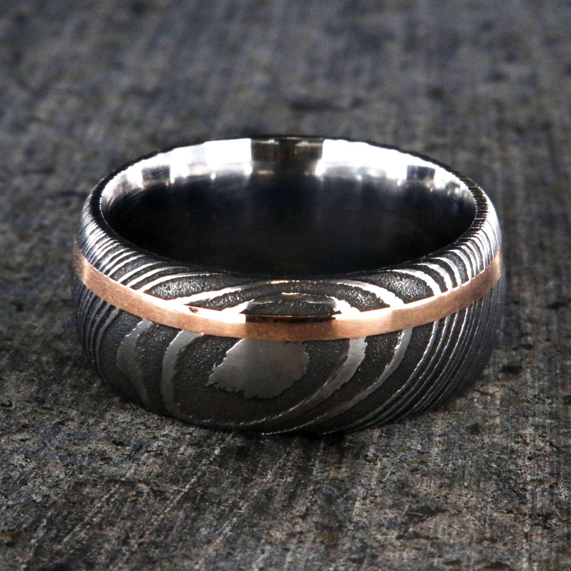 10mm wide black Damascus steel wedding band for men with two-tone pattern, an off-center rose gold inlay, and a polished inside