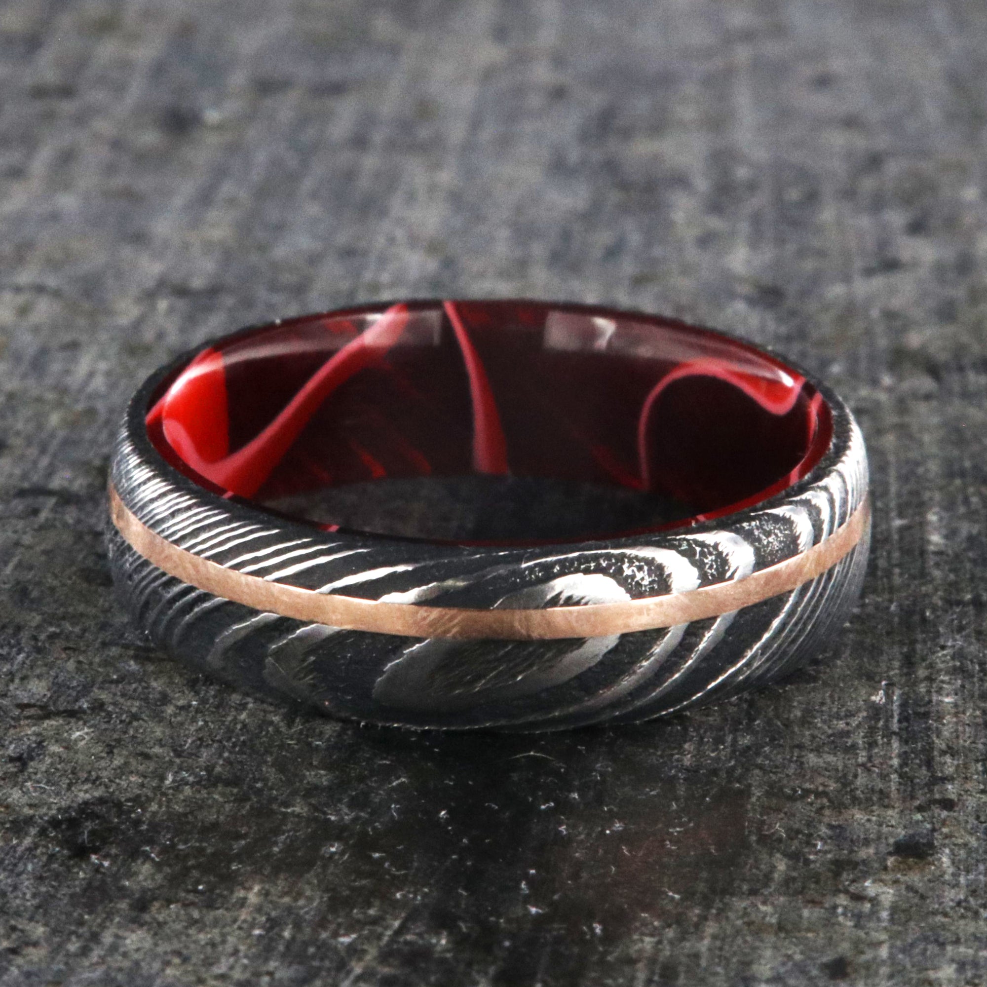 6mm wide black Damascus steel ring with a thin off-centered rose gold inlay and dark and bright red swirled acrylic sleeve