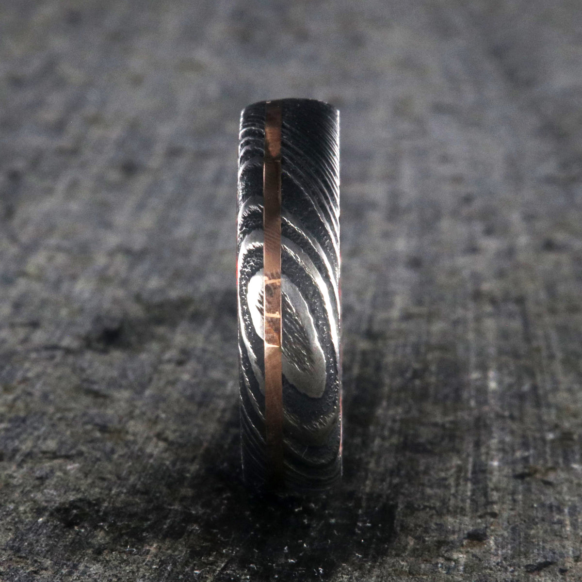 6mm wide black Damascus steel ring with a thin off-centered rose gold inlay and dark and bright red swirled acrylic sleeve