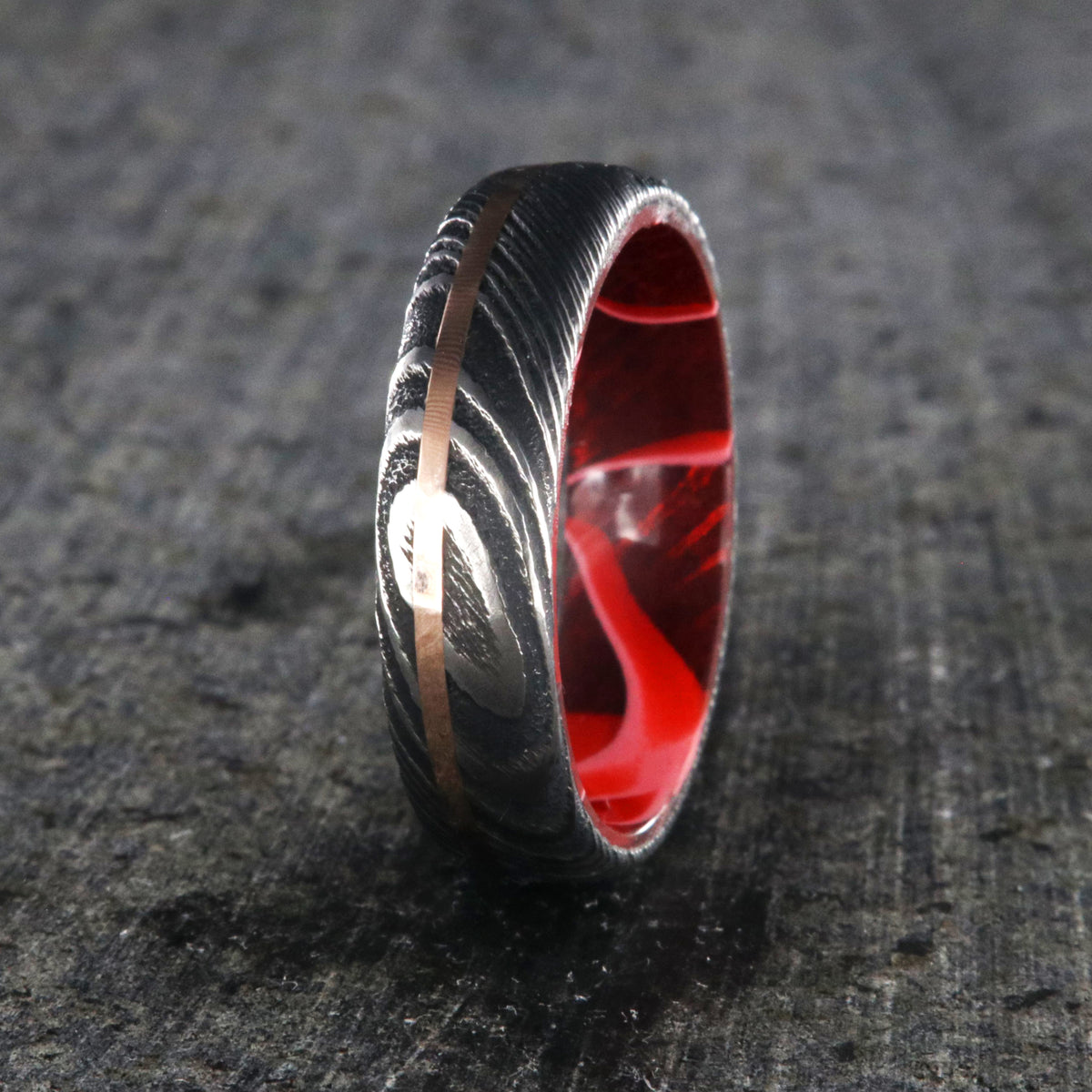6mm wide black Damascus steel ring with a thin off-centered rose gold inlay and dark and bright red swirled acrylic sleeve