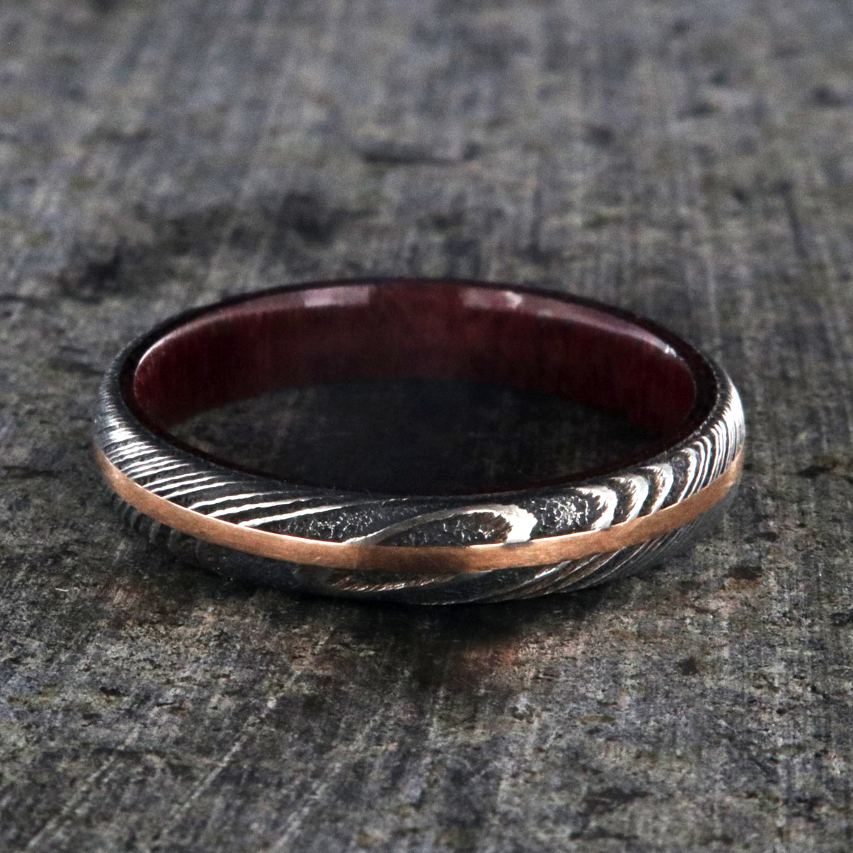 4mm wide women&#39;s wedding band made with black Damascus steel with a centered rose gold inlay and purple rain wood sleeve