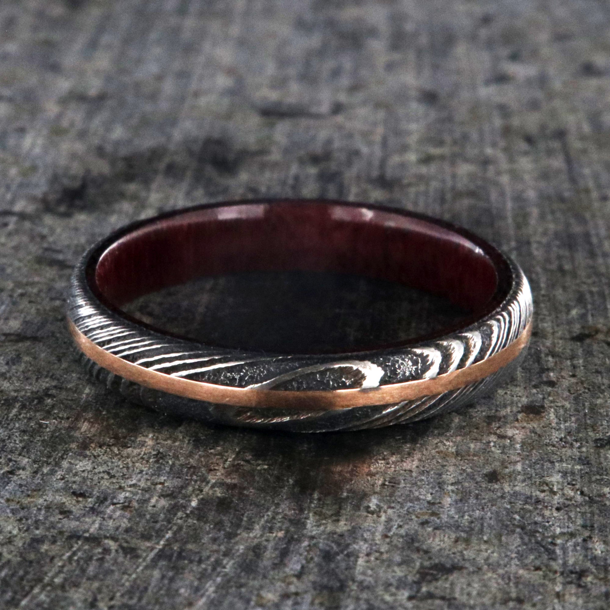 4mm wide women's wedding band made with black Damascus steel with a centered rose gold inlay and purple rain wood sleeve