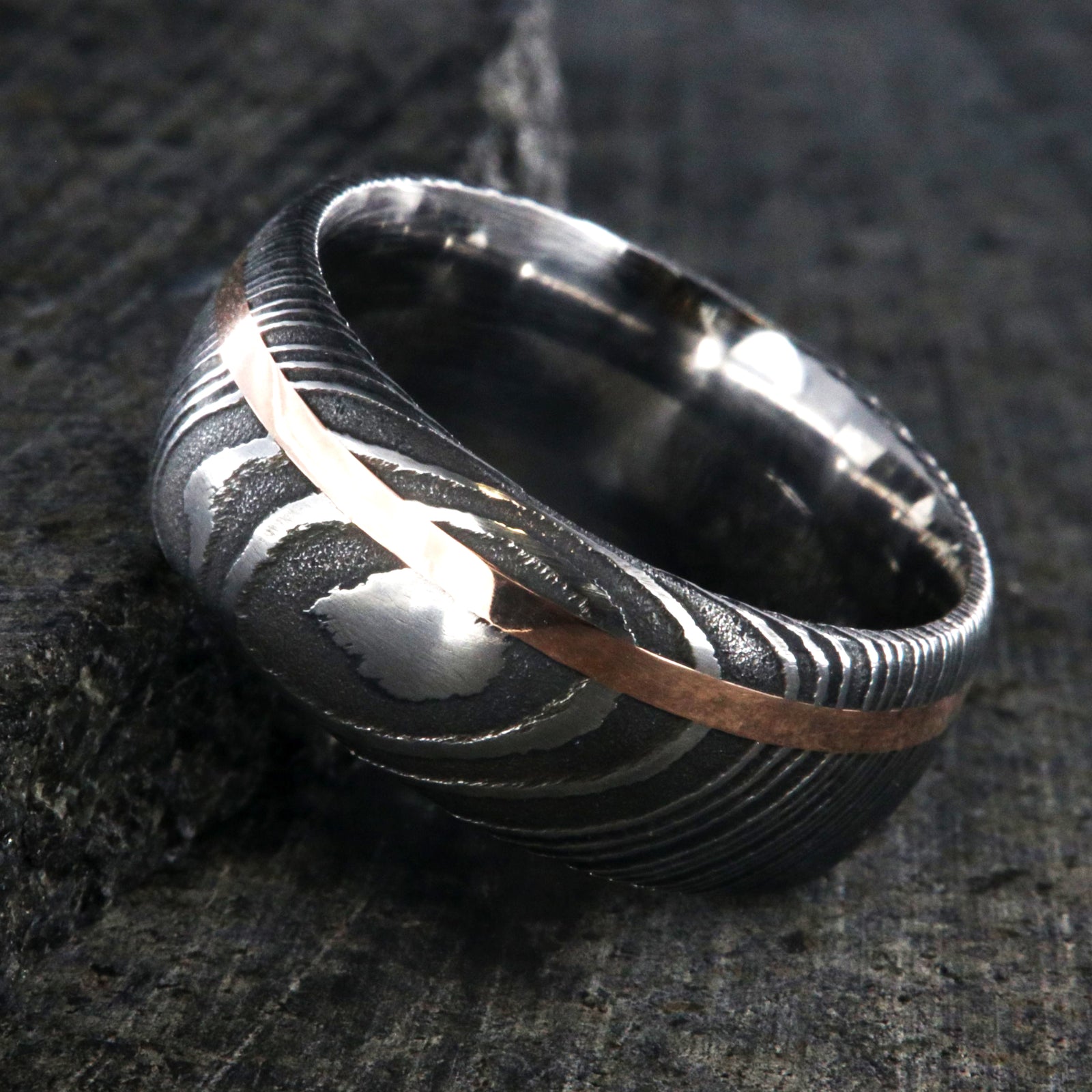 10mm wide black Damascus steel wedding band for men with two-tone pattern, an off-center rose gold inlay, and a polished inside