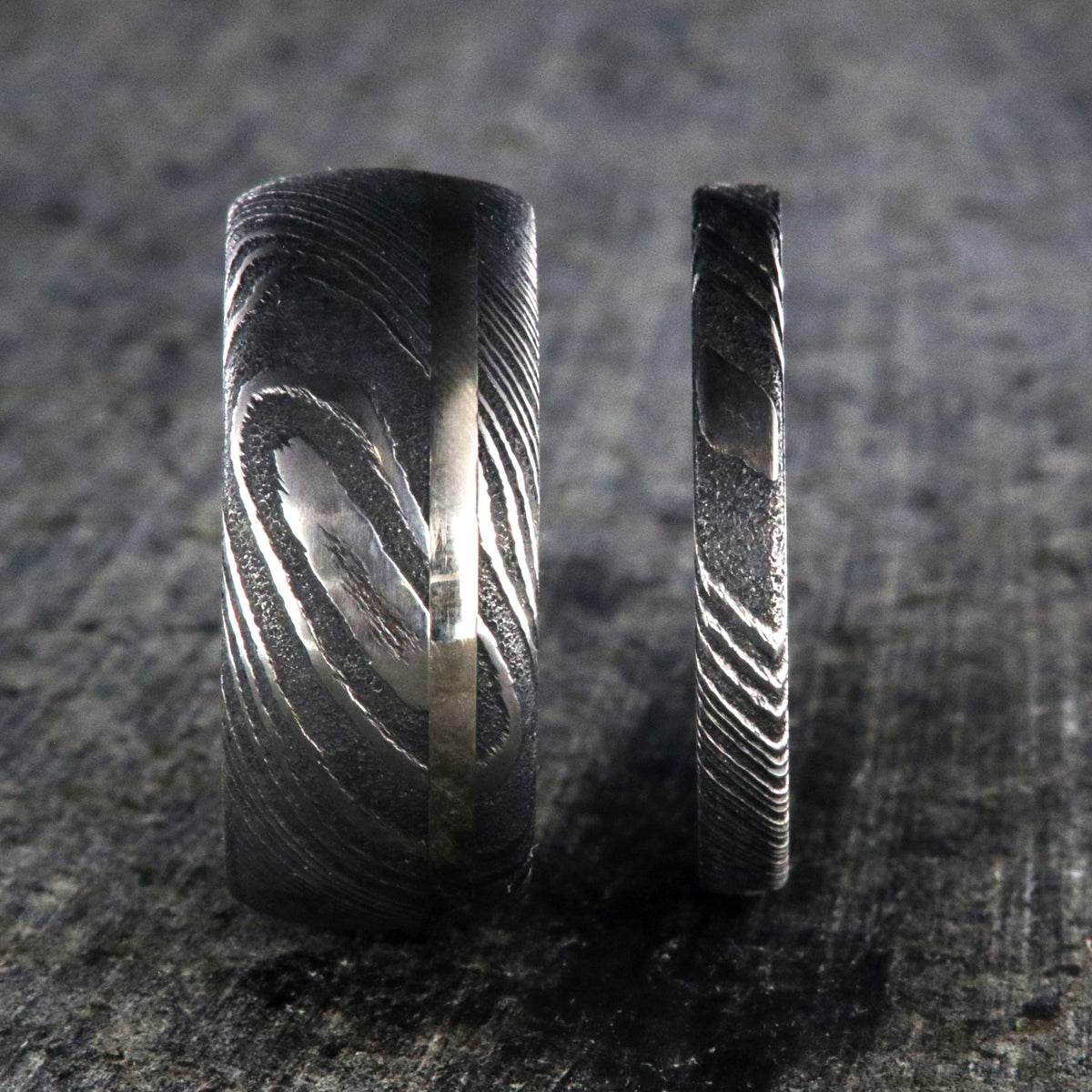 10mm wide men&#39;s Damascus steel wedding band with off-center white gold inlay with a 3mm wide women&#39;s wedding band of Damascus steel