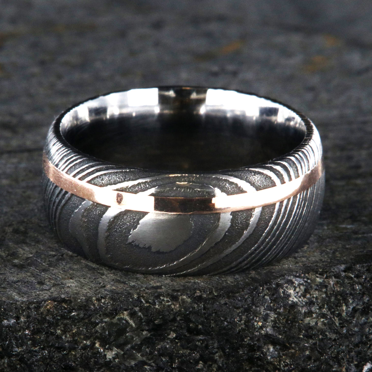 10mm wide black Damascus steel wedding band for men with two-tone pattern, an off-center rose gold inlay, and a polished inside