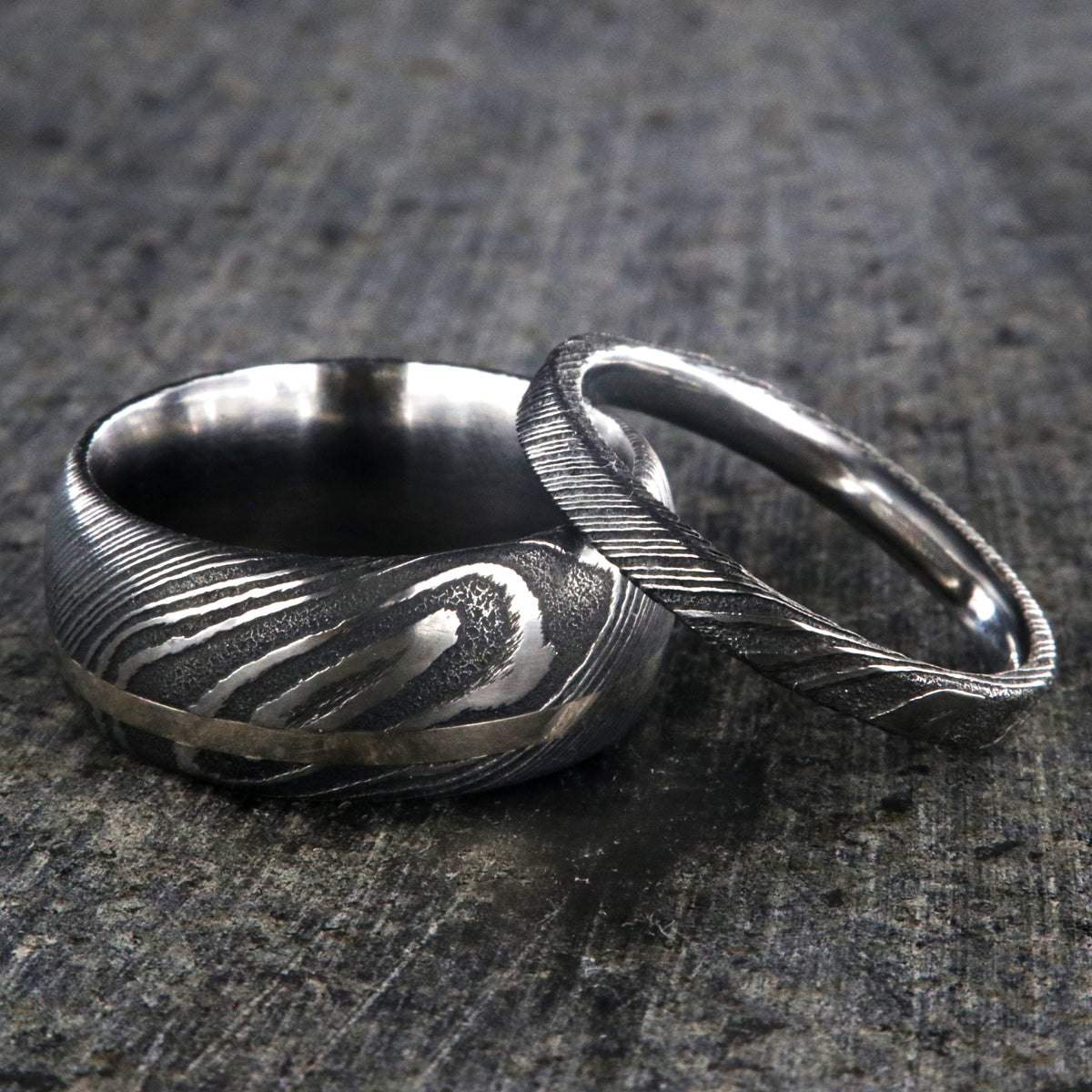 10mm wide men&#39;s Damascus steel wedding band with off-center white gold inlay with a 3mm wide women&#39;s wedding band of Damascus steel