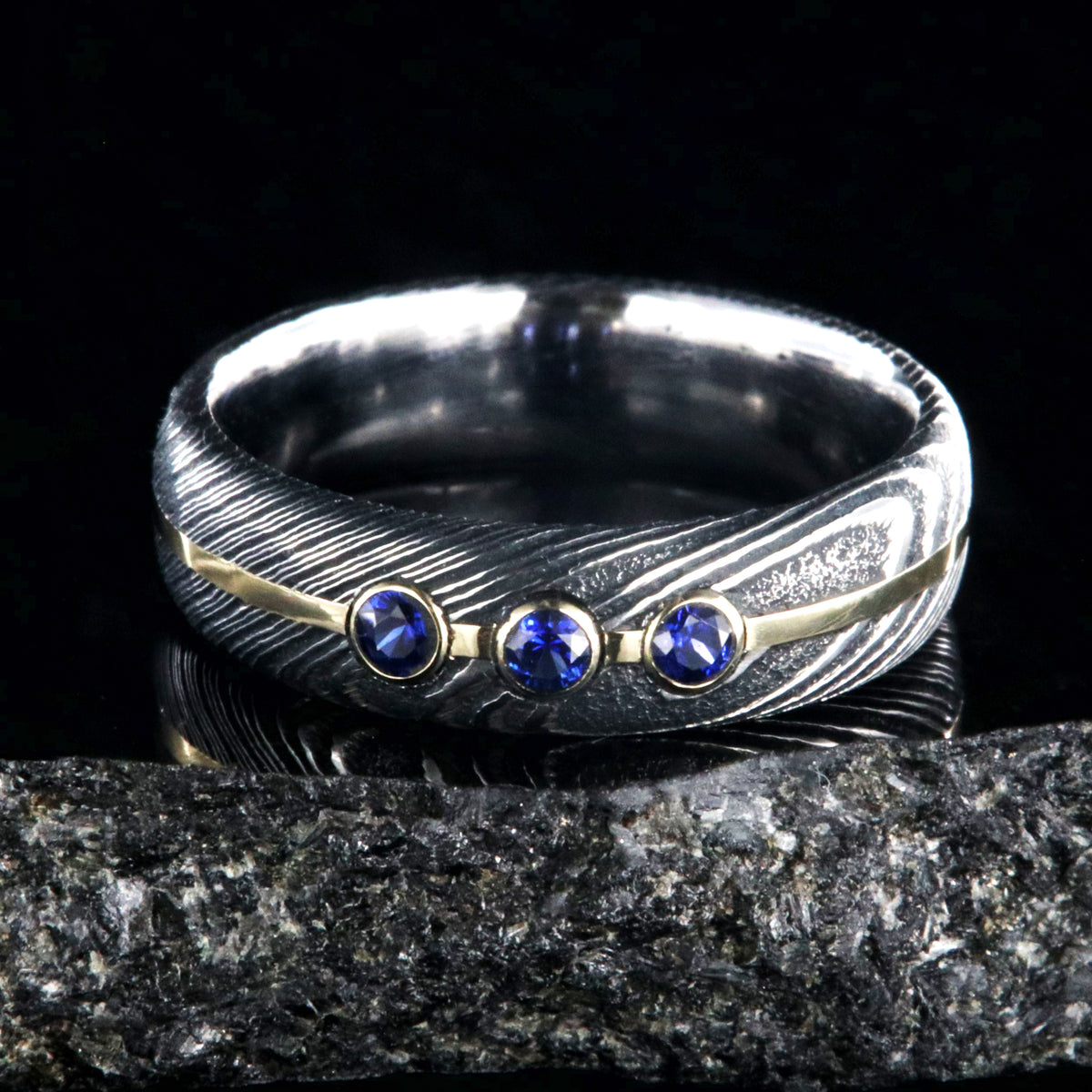 6mm wide black Damascus steel wedding band with 3 blue sapphire stone with a yellow gold center inlay