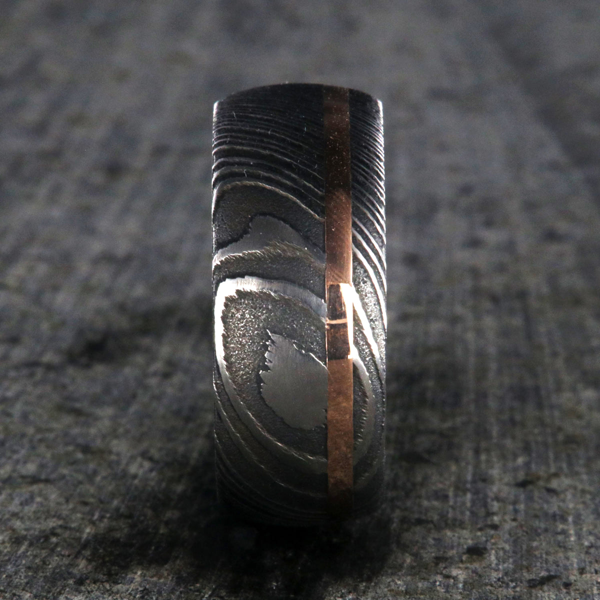 10mm wide black Damascus steel wedding band for men with two-tone pattern, an off-center rose gold inlay, and a polished inside