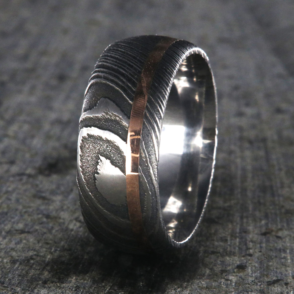 10mm wide black Damascus steel wedding band for men with two-tone pattern, an off-center rose gold inlay, and a polished inside