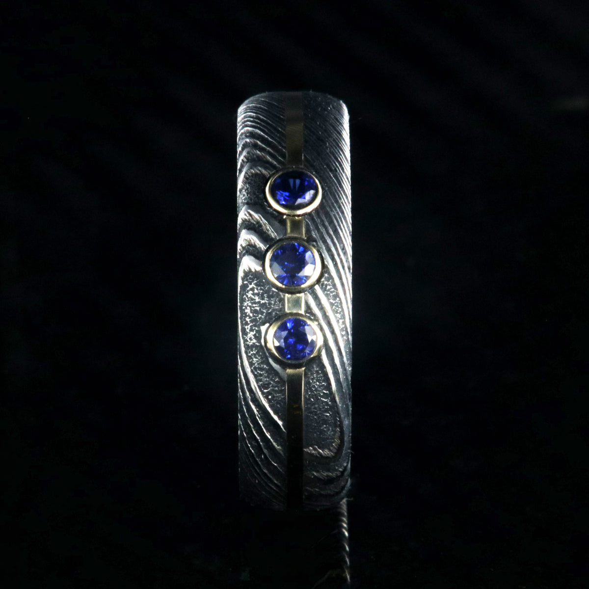 6mm wide black Damascus steel wedding band with 3 blue sapphire stone with a yellow gold center inlay