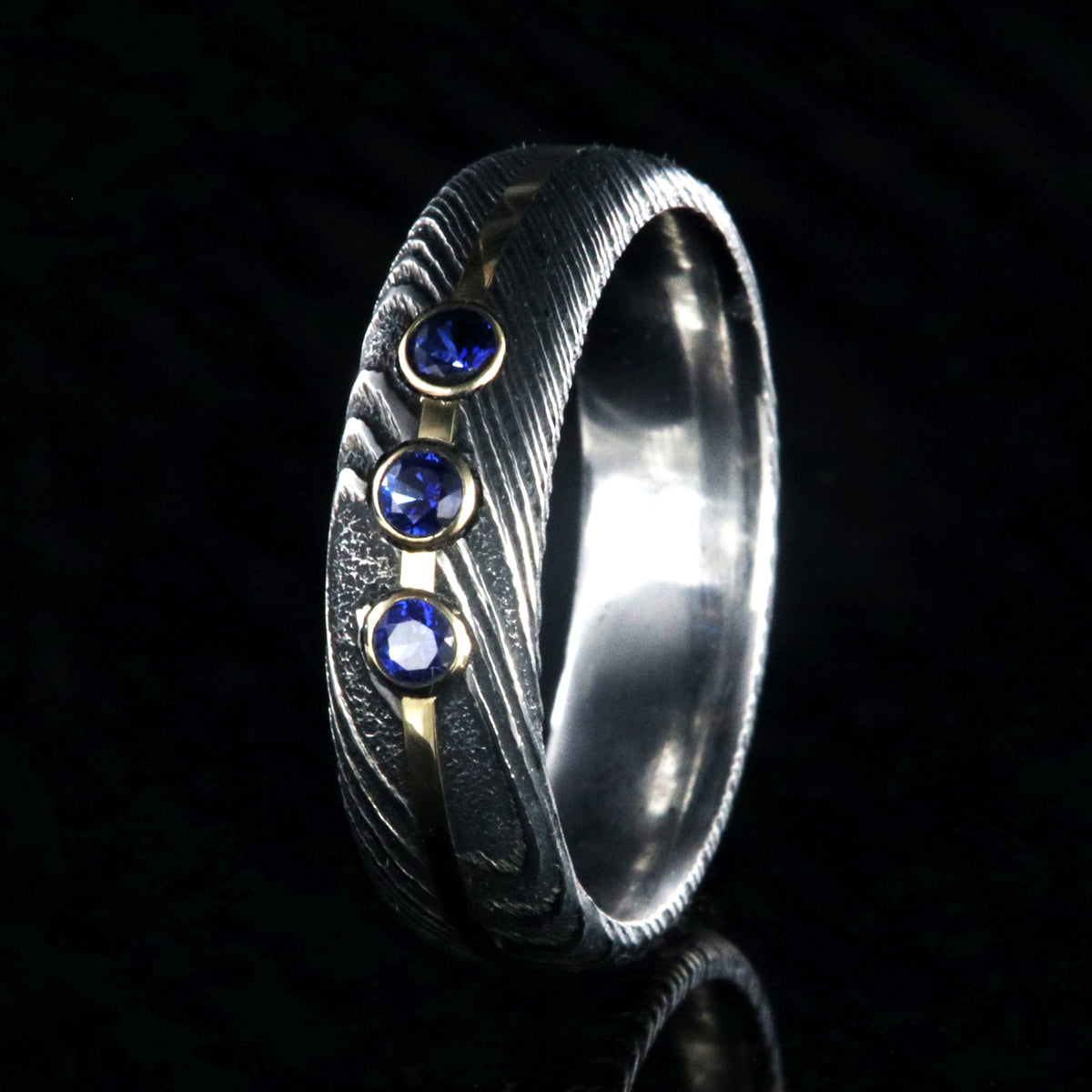 6mm wide black Damascus steel wedding band with 3 blue sapphire stone with a yellow gold center inlay