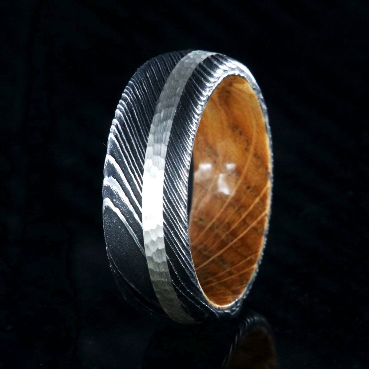 8mm wide black Damascus steel wedding band with a hammered 2mm wide sterling silver inlay and a whiskey barrel sleeve