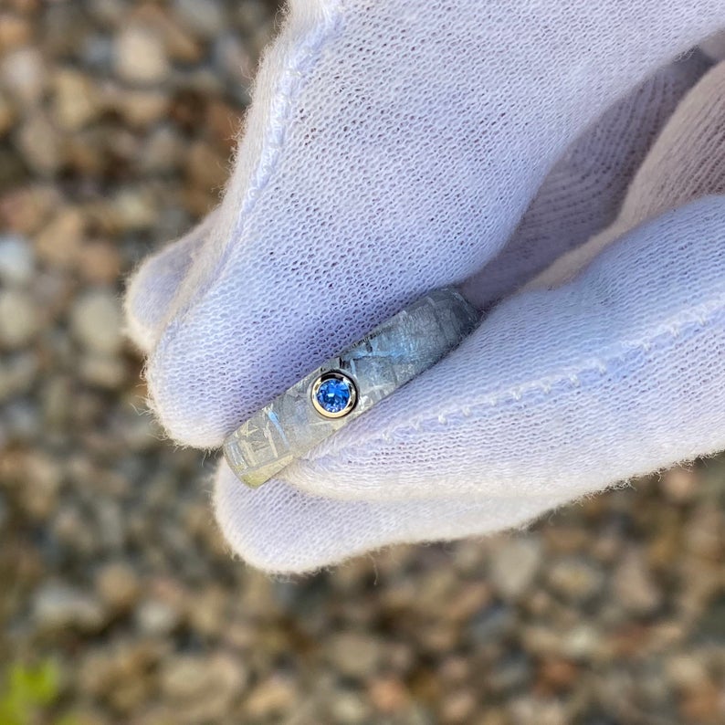 5mm wide women&#39;s meteorite ring with rounded profile and a blue sapphire set in a gold bezel held by a white gloved hand