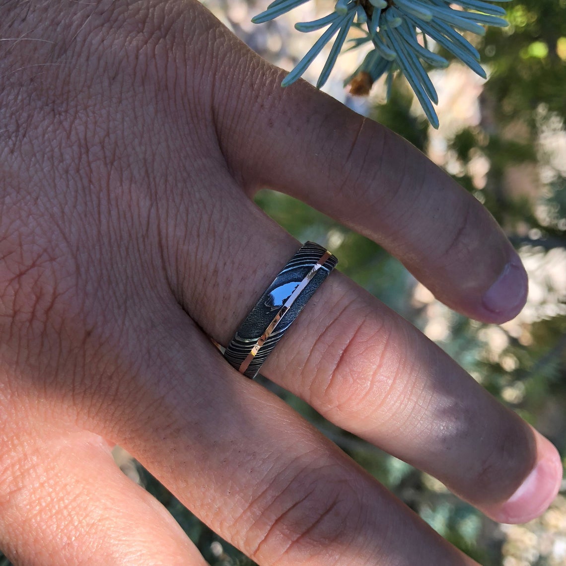 6mm wide black Damascus steel ring with an off-center white gold inlay on a man&#39;s hand