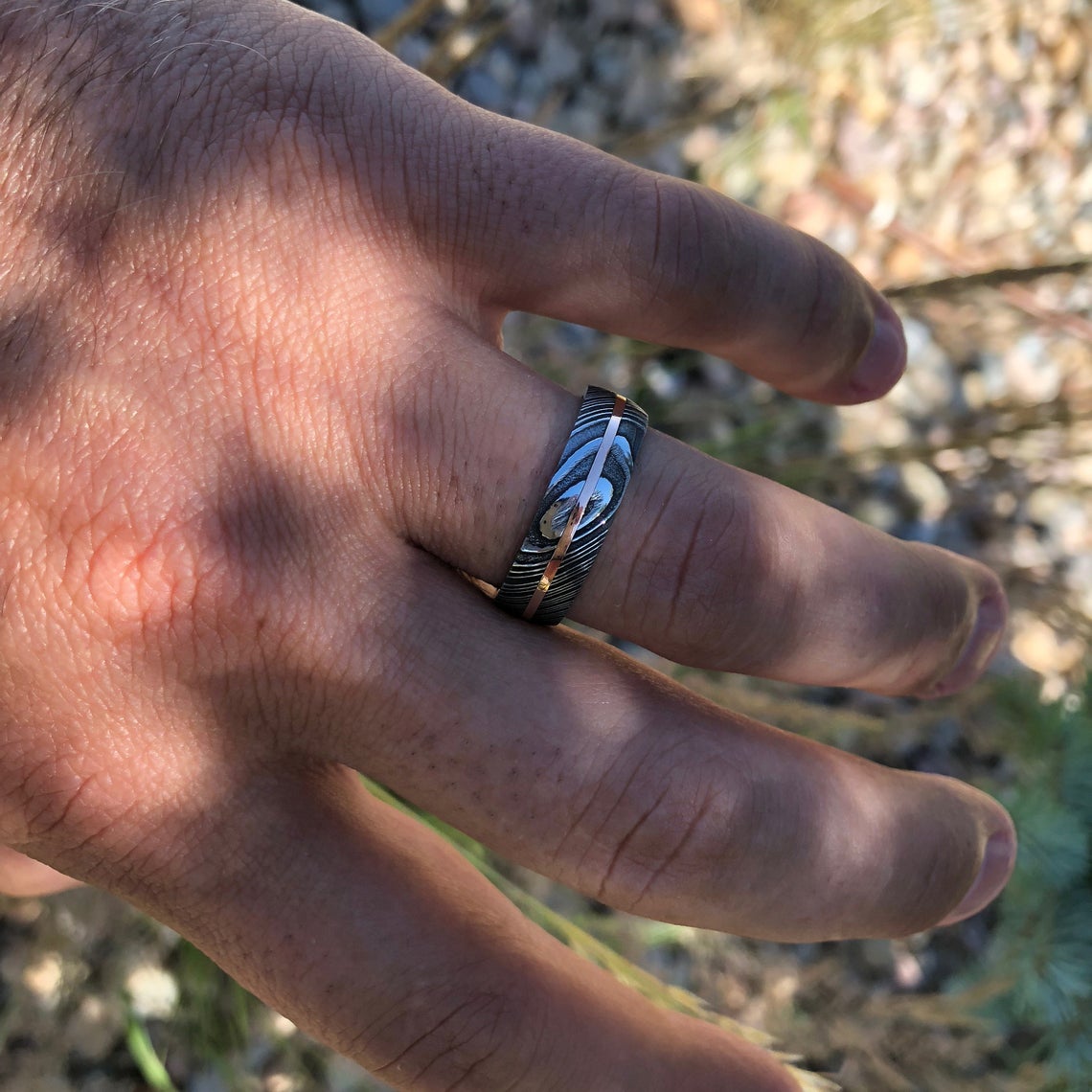 7mm wide men&#39;s Damascus steel wedding ring with a thin centered rose gold inlay and Arizona ironwood sleeve on man&#39;s hand