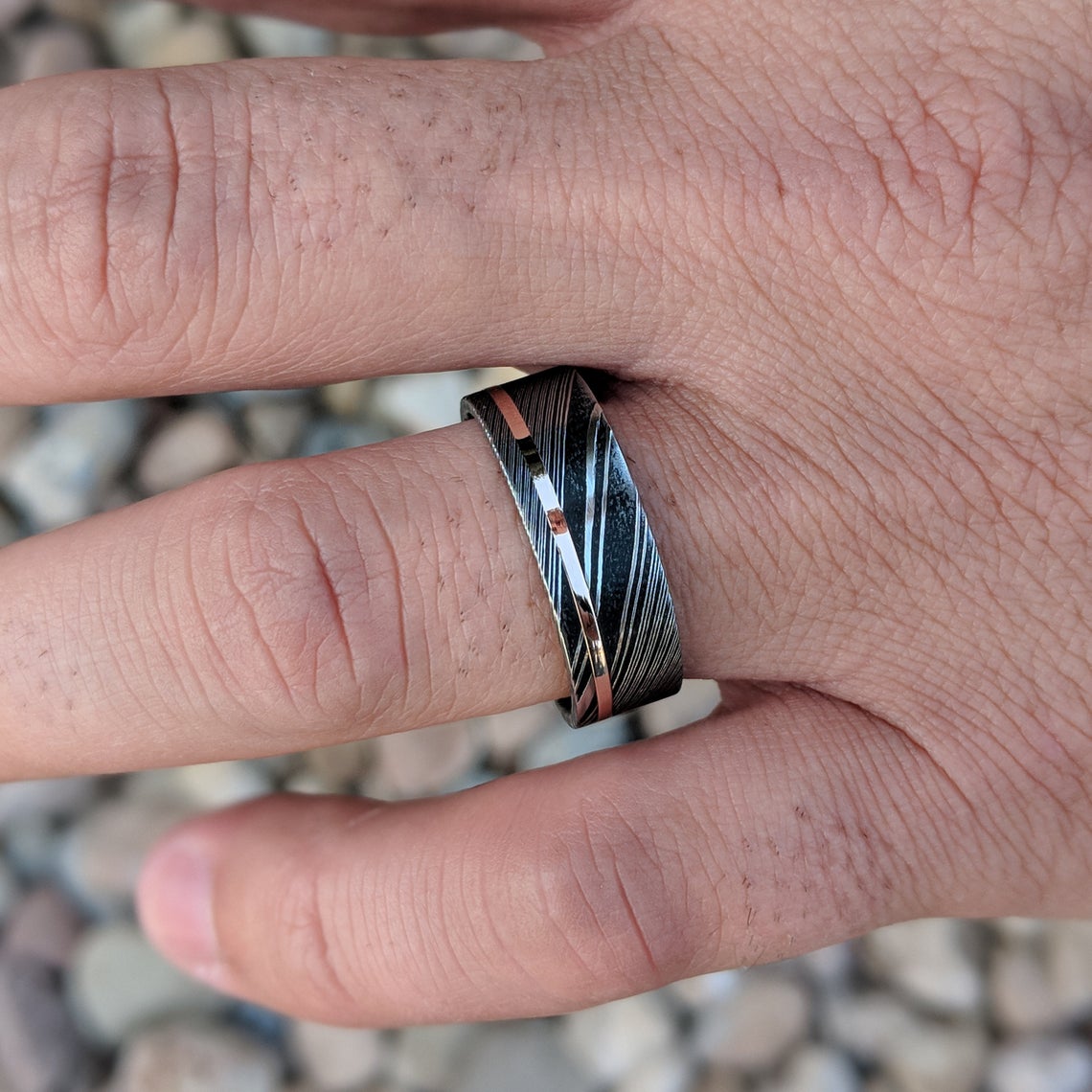 8mm wide black Damascus steel ring with a flat profile, polished inside, and a thin off-centered rose gold inlay on man&#39;s hand