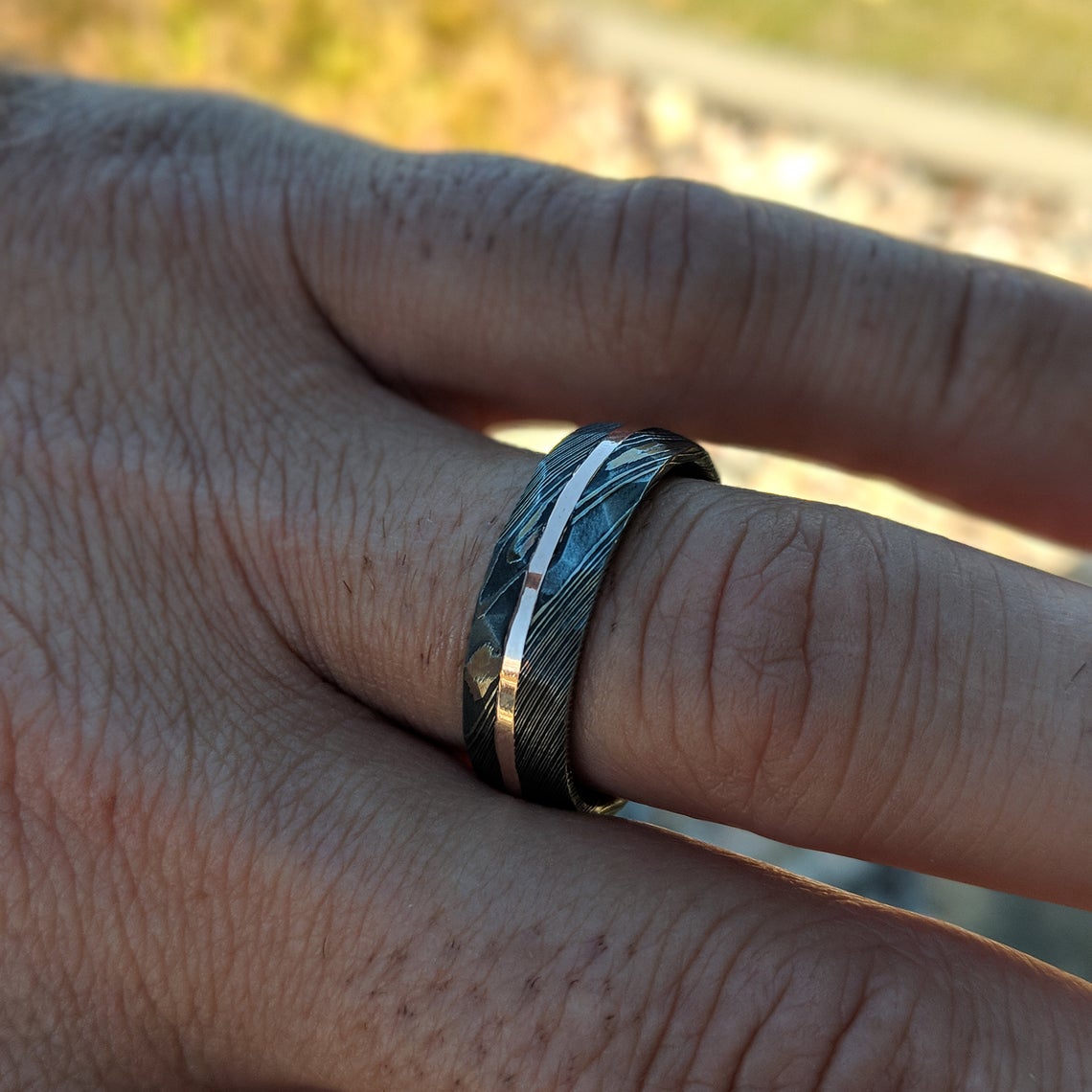 8mm wide black Damascus steel ring with two-tone pattern with a centered rose gold inlay and a big hammered finish on a man&#39;s hand
