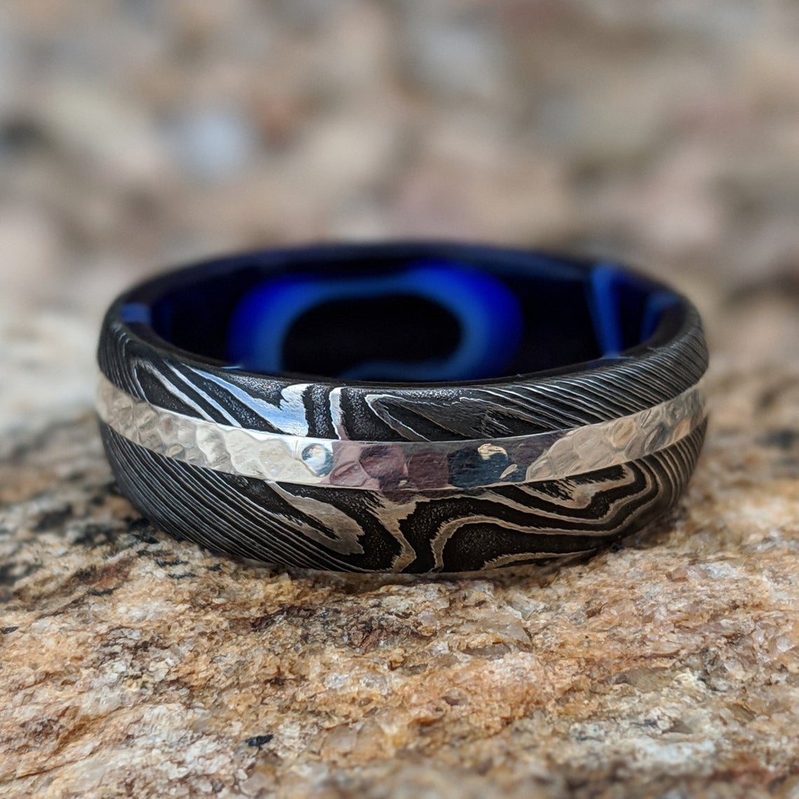 8mm wide black Damascus steel wedding band with a 2mm wide white gold inlay with a dark blue acrylic sleeve