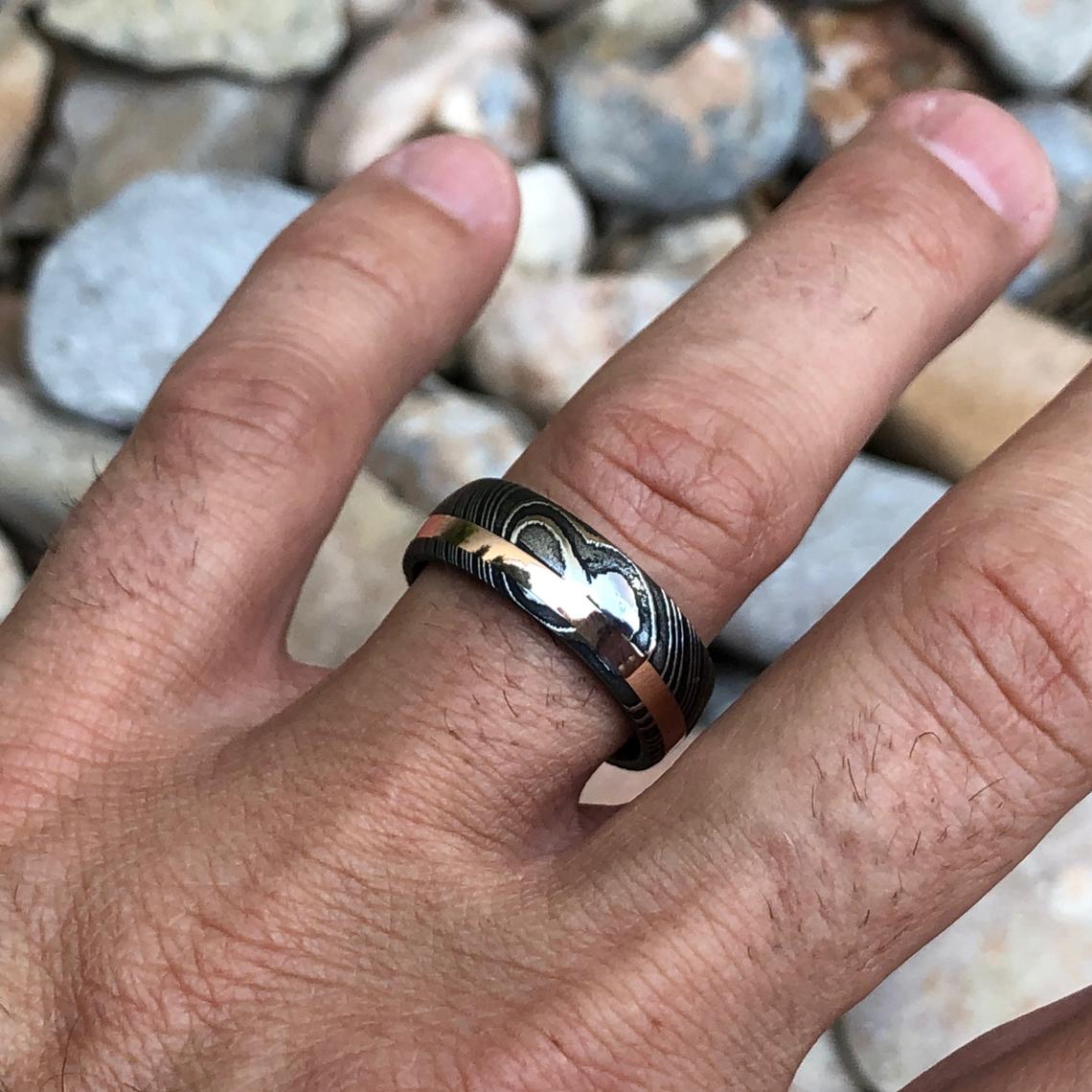 8mm wide black Damascus steel wedding band with a 2mm wide rose gold inlay and rounded profile on a man&#39;s hand