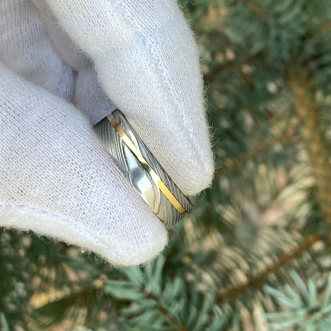 6mm wide white Damascus steel ring with a 14k yellow gold off-center inlay and sleeve held by a gloved hand