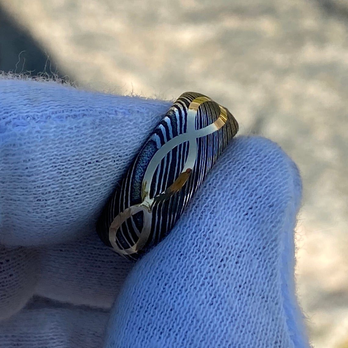 8mm wide Black Damascus steel ring with a 14k yellow gold infinity inlay held by a white glove