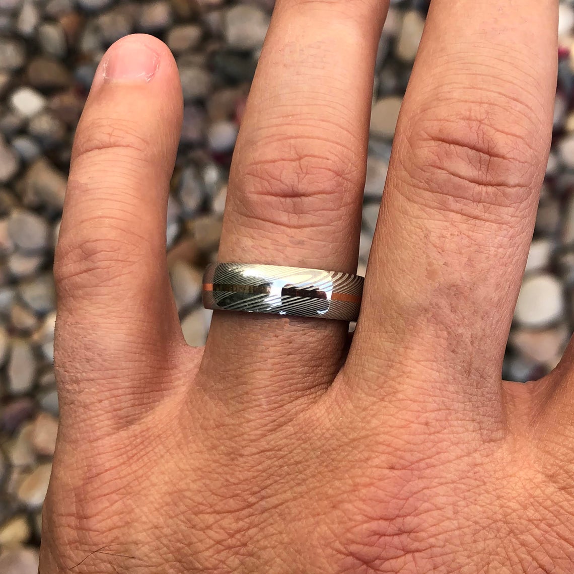 7mm wide white Damascus steel ring with a centered rose gold inlay on man&#39;s hand