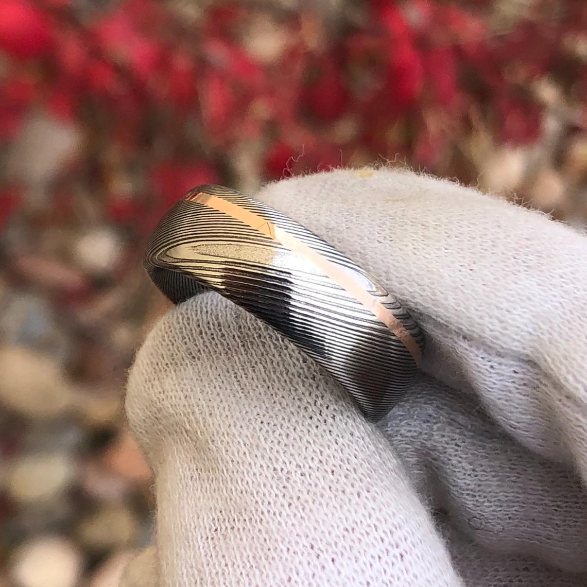 8mm wide Damascus steel ring with an off-centered  rose gold inlay held by a white glove